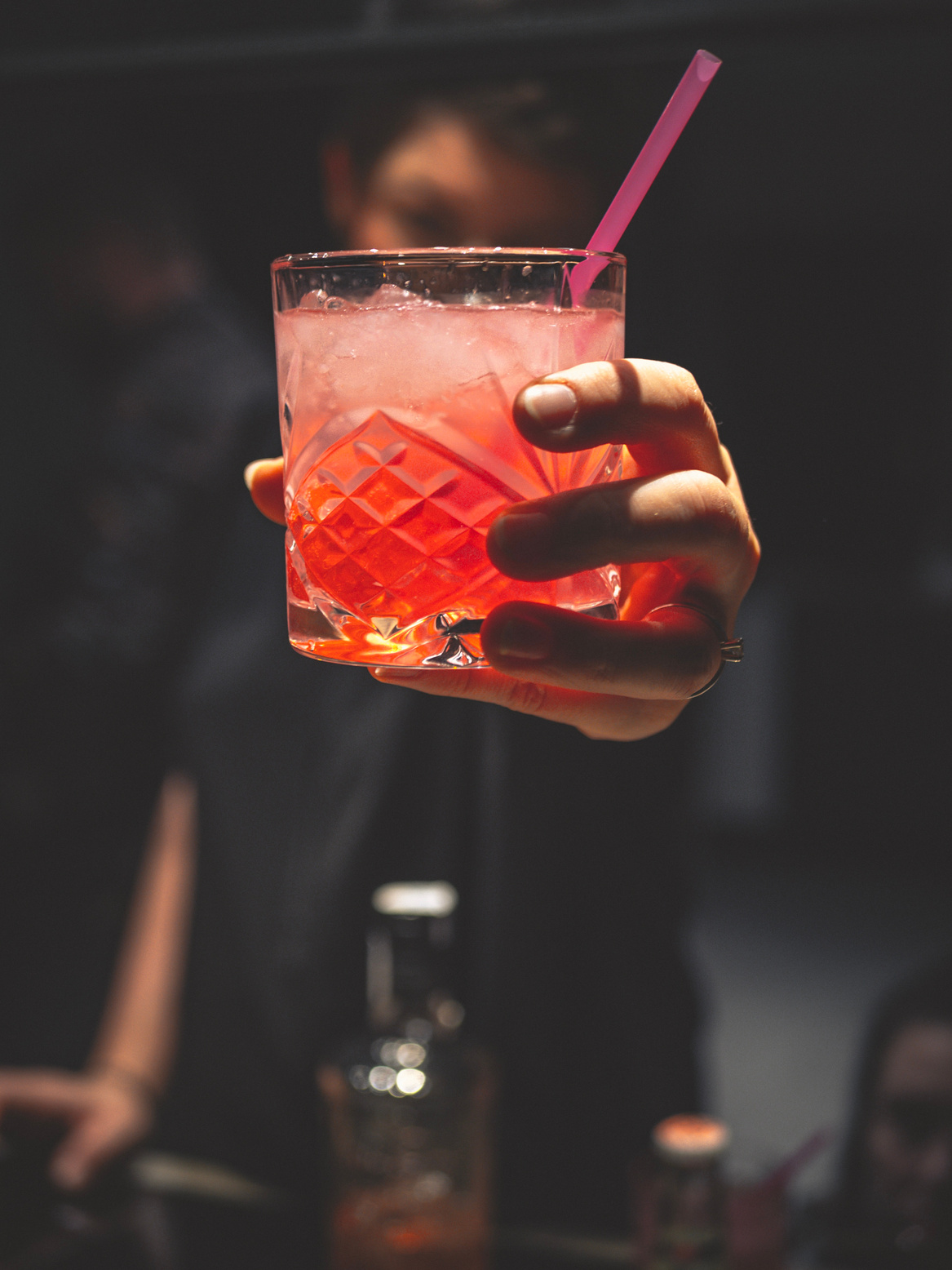 Man Holding Forward A Glass Of Iced Pinkish Colored Cocktail With A Straw